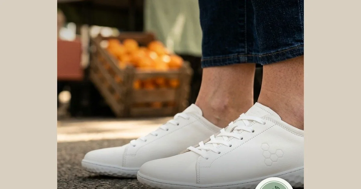 Persona de pie con las Vivobarefoot Geo Court III blancas, las dos zapatillas completas a la vista, mercado de fondo.